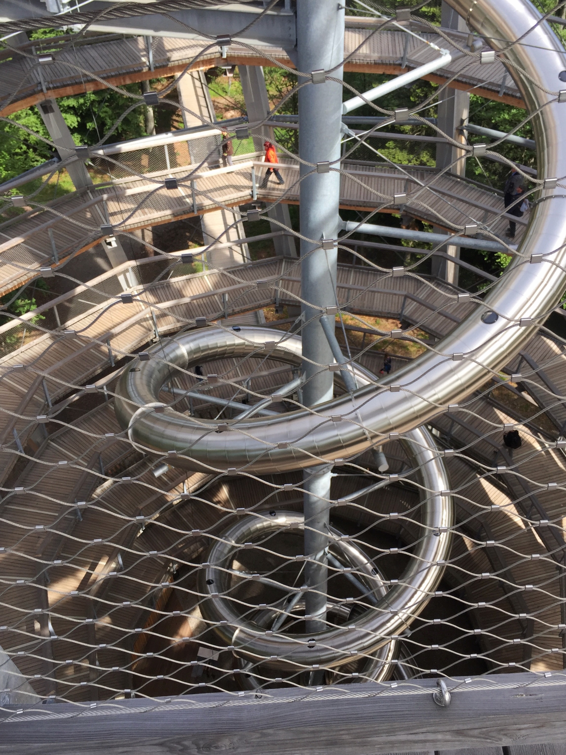 Giant metal spiral slide with surrounding wooden walkway ramp, viewed from above through a protective chain-link fence