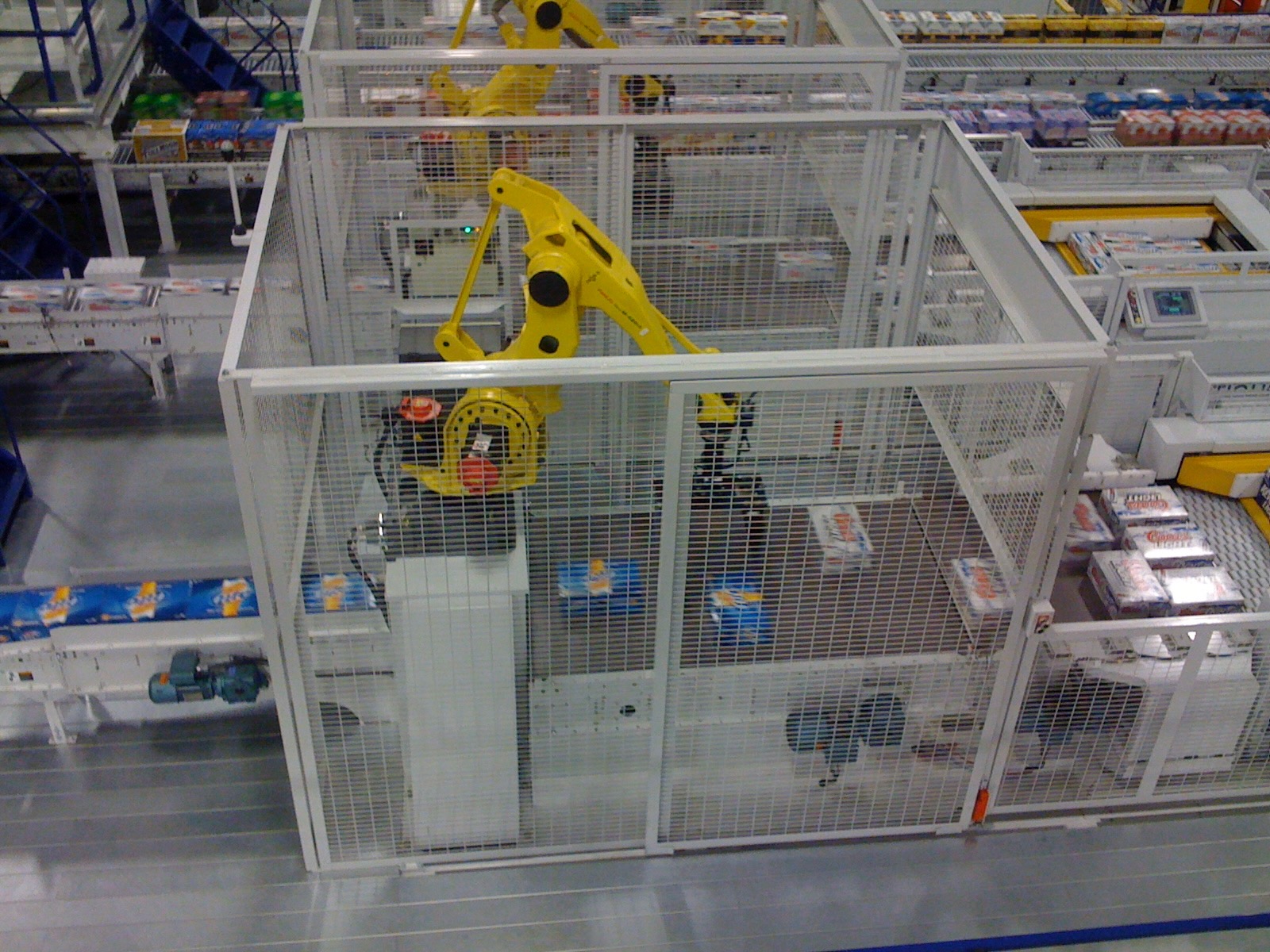 A yellow industrial articulated robot arm (FANUC-style) enclosed in a wire-mesh safety cage, positioned within a beverage packaging/bottling facility. Conveyor belts carrying cases of canned drinks run through the surrounding warehouse, with stacked product visible on shelving in the background and a concrete factory floor.